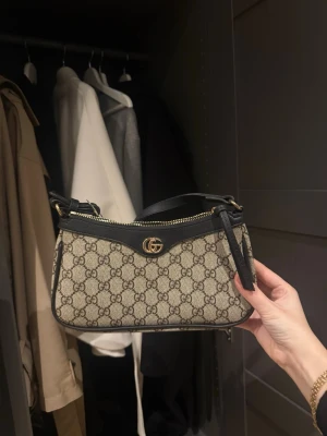  Supreme axelremsväska - Snygg axelremsväska Supreme-mönster. Väskan är beige med svarta läderdetaljer och guldfärgad logga framtill. Praktisk dragkedja upptill och stilren form som passar till många outfits.