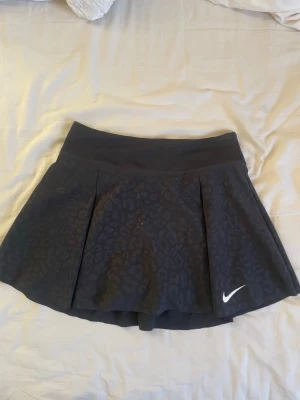 Svart kjol med inbyggda shorts- Nike - Svart träningskjol med inbyggda shorts i XS. Den har ett svagt leopardmönster och är väldigt skön. Den har en ficka för telefon i shortsen.