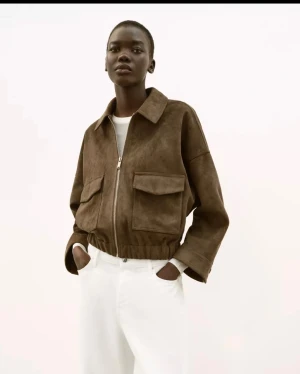 Brun mockajacka med dragkedja - Helt ny med lappar kvar! Snygg brun jacka i mockaimitation med dragkedja framtill, två stora fickor på bröstet och elastisk midja. Jackan har avslappnad passform, klassisk krage och raka ärmar med knappdetalj vid ärmslut. Perfekt för en trendig och stilren look. Aldrig använd! 