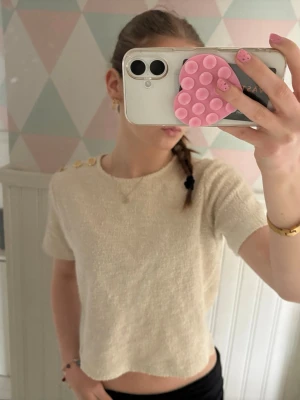 Krämvit topp med guldknappar - Trendig krämvit topp med korta ärmar och tre dekorativa guldfärgade knappar på axeln. Tillverkad i ett mjukt, strukturerat material som ger en lyxig känsla. ☺️☺️