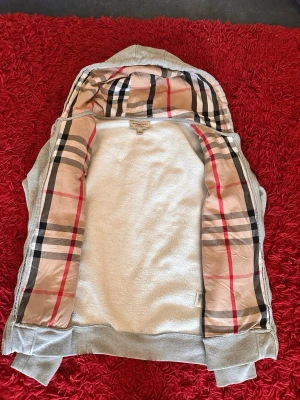 Grå hoodie från Burberry med dragkedja - Snygg grå hoodie från Burberry med dragkedja och klassiskt rutigt foder i huvan och längs insidan. Tröjan har dragsko i huvan, ribbade muddar och fickor framtill. Perfekt för en stilren och avslappnad look.