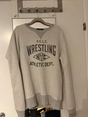 Grå sweatshirt från Polo Ralph Lauren - Snygg grå sweatshirt från Polo Ralph Lauren med tryck på bröstet där det står 'Wrestling Athletic Dept.'. Tröjan har rund halsringning, ribbade muddar och avslappnad passform. Perfekt för en sportig och avslappnad stil. Storlek m| skick 9,5/10|