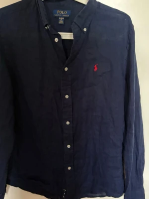 Mörkblå linneskjorta från Polo Ralph Lauren - Snygg mörkblå skjorta i linne från Polo Ralph Lauren med klassisk krage, knappar framtill och ikonisk röd logga broderad på bröstet. Långärmad och perfekt för en stilren look. Den är storlek 18/20 barn men passar typ S vuxen.