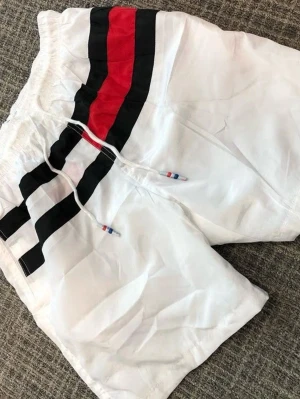 Vita shorts med röd och svart rand - Säljer ett par vita shorts med breda svarta och en röd rand längs sidan. Shortsen har snörning i midjan med små färgdetaljer på snörena. Perfekta för sommaren och har en sportig vibe.