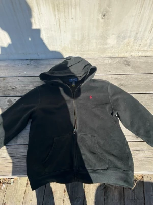 Svart hoodie från Polo Ralph Lauren - Svart hoodie från Polo Ralph Lauren med dragkedja och huva. Klassisk design med röd broderad logga på bröstet och två fickor framtill. Mjuk insida och ribbade muddar. Perfekt för en avslappnad och stilren look.