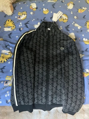 Svart mönstrad track jacket Lacoste - Svart track jacket från Lacoste med grått geometriskt mönster och klassisk grön krokodillogga på bröstet. Helt sprillans ny med tags fortfarande på!
