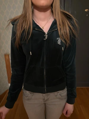 Juicy Couture svart velour hoodie - Mörkgrön velour hoodie från Juicy Couture med dragkedja, huva och fickor framtill. Stora strasstenar bildar texten 'Juicy' på ryggen och ett emblem på bröstet. Perfekt för en chill och trendig look.