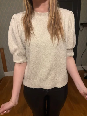 Beige stickad tröja med puffärm - Trendig beige stickad tröja med puffärmar och ribbade muddar. Tröjan har rund halsringning och är tillverkad i ett mjukt, strukturerat material. Perfekt för dig som gillar en enkel men snygg look.