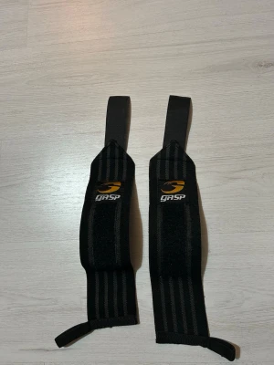 Svarta wristwraps från GASP - Ett par svarta handledslindor från GASP med gul logga. Tillverkade i elastiskt material med kardborreband och tumögla för extra stöd vid styrketräning. Perfekta för gymmet när du vill ha stabilitet och support.