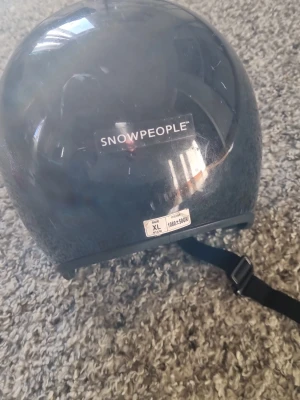 Svart hjälm från Snowpeople XL - Säljer en svart hjälm från Snowpeople i storlek XL. Hjälmen har justerbart hakband och vadderad insida för extra komfort. Passar perfekt för vintersport eller annan aktivitet där hjälm behövs. En knapp är trasig