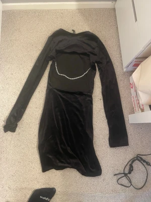 Svart sammetsklänning med strassdetalj - Svart sammetsklänning med lång ärm och en snygg strassdetalj över magen. Klänningen är kort och har en figurnära passform som ger en cool och elegant look. Perfekt för dig som vill sticka ut med en unik detalj.
