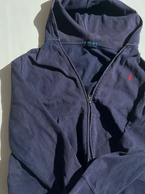 Mörkblå hoodie från Polo Ralph Lauren XS/S - Mörkblå hoodie från Polo Ralph Lauren med dragkedja och klassisk huva. Liten röd logga broderad på bröstet. Tillverkad i mjukt bomullsmaterial, perfekt för en avslappnad och stilren look.