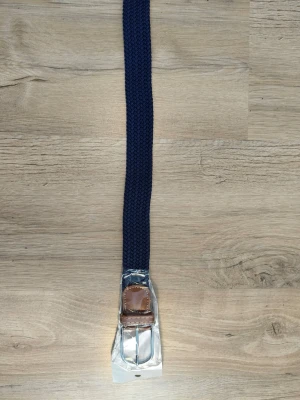 Mörkblå flätat bälte med läderdetalj - Snyggt mörkblått flätat bälte med bruna läderdetaljer och silvrigt spänne. Perfekt accessoar för att lyfta din outfit med en klassisk touch. Bältet har en stilren design och passar till många olika looks.