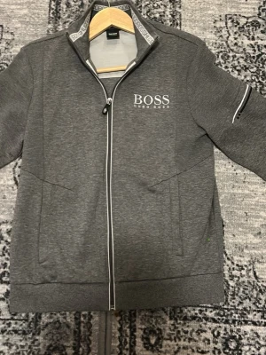 Grå zip-hoodie från Hugo Boss HELY NY - Snygg grå hoodie från Hugo Boss med dragkedja framtill och vit logga på bröstet. Tröjan har ståkrage, två sidofickor och detaljerade sömmar samt en diskret ficka med dragkedja på ena ärmen. Perfekt för en stilren och sportig look.