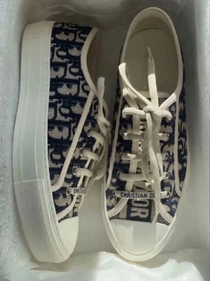 Christian Dior sneakers med monogram - Snygga sneakers från Christian Dior med blått och vitt monogrammönster, vita snören och tjock vit sula. Ovandelen är i textil med tydlig Dior-logga på plösen och sidan. Perfekta för dig som vill sticka ut med exklusiv streetstyle.