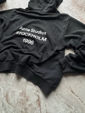 Svart hoodie Acne Studios Stockholm - Svart hoodie från Acne Studios med vit text på ryggen där det står 'Acne Studios Stockholm 1996'. Klassisk passform med huva och magficka. Mjuk bomullskvalitet och ribbade muddar vid ärmslut och nederkant. Perfekt för en stilren och avslappnad look.
