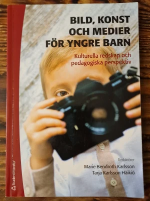 Bild, konst och medier för yngre barn - Kursbok med titeln 'Bild, konst och medier för yngre barn', redaktörer Marie Bendroth Karlsson och Tarja Karlsson Häikiö. Boken har ett mjukt omslag i rött och vitt med ett foto på ett barn. Perfekt för dig som pluggar pedagogik, bild eller barnkultur.