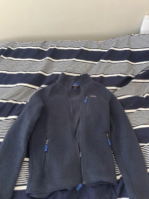 Mörkblå fleecejacka från Patagonia - Mysig mörkblå fleecejacka från Patagonia med dragkedja framtill och tre fickor med blå detaljer. Jackan har hög krage och är perfekt för lager-på-lager. Materialet är mjukt och värmande, perfekt nu för våren och kyliga sommarkvällar. Storlek Medium