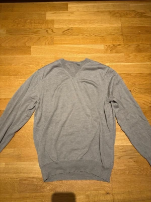 Ljusgrå  merinoulltröja - Snygg ljusgrå stickad tröja i extra fin merinoull. Tröjan har V-ringning, långa ärmar och ribbade muddar vid ärmslut och nederkant. Perfekt för dig som gillar stilrena och bekväma plagg i mjukt material. Strl L