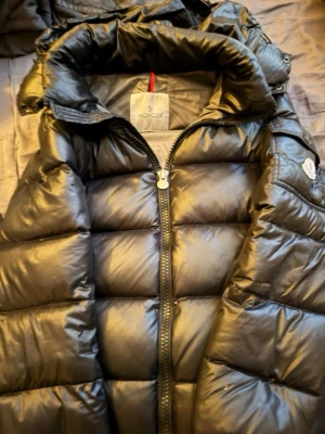 Svart dunväst från Moncler - Snygg svart dunväst från Moncler med hög krage och quiltad design. Västen har dragkedja framtill och en ficka med Moncler-logga på bröstet. Tillverkad i glansigt material som ger en exklusiv känsla. Perfekt för lager-på-lager och streetwear. Det står storlek 4