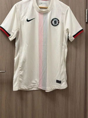Chelsea vit matchtröja Nike - Chelsea FC:s vita matchtröja från Nike med svarta och röda ränder längs framsidan. Tröjan har klubbmärke på bröstet, svart Nike-logga och detaljer i svart och rött på ärmsluten. Perfekt för fotboll eller träning.