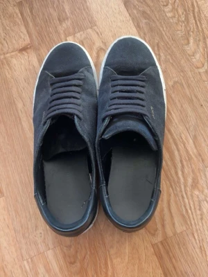 Svarta sneakers från Axel Arigato - Snygga svarta sneakers från Axel Arigato med klassisk låg profil och vit sula. Skorna har ovandel i mocka och textil, svarta skosnören och diskret logga i guld på sidan. Perfekta för en stilren och avslappnad look.