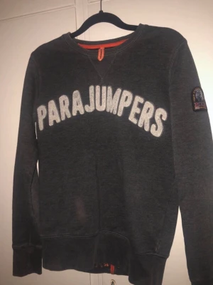 Mörkgrå sweatshirt från Parajumpers - Mörkgrå sweatshirt från Parajumpers med stort vitt tryck över bröstet och klassisk logotyp-patch på ärmen. Tröjan har rund halsringning, ribbade muddar och en avslappnad passform. Perfekt för en cool och avslappnad stil.