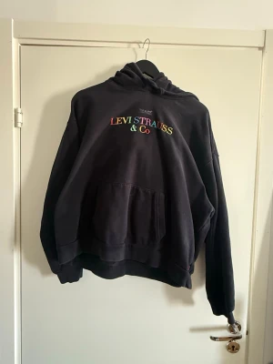 Svart hoodie från Levi Strauss & Co - Svart hoodie med broderad logga i regnbågsfärger från Levi Strauss & Co. Klassisk känguruficka framtill och justerbar huva med dragsko. Mjuk bomullskänsla och avslappnad passform, perfekt för en chill och stilren look. XL/L