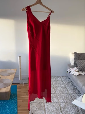 Röd långklänning med asymmetrisk fåll - Snygg röd långklänning med smala axelband och v-ringning. Klänningen har en asymmetrisk, något transparent fåll som ger en cool och modern look. Perfekt för dig som vill sticka ut med en elegant och enkel stil.