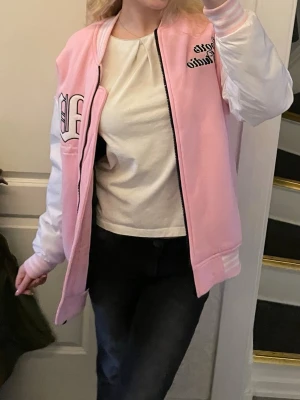 Ljusrosa cool vårjacka med detaljer - Säljer en snygg bomberjacka i rosa med vita ärmar och svarta detaljer. Jackan har broderade bokstäver på bröstet och dragkedja framtill. Ribbstickade muddar vid ärmslut och nederkant. Perfekt för dig som vill ha en trendig och sportig look. S. 