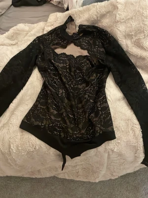 Svart spetsbody med lång ärm - Snygg svart body i spets med lång ärm och hög hals. Transparent spets med blommönster och en unik cut-out detalj vid halsen. Perfekt för dig som vill ha något extra till outfiten. Ingen synlig logga eller märke.