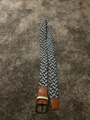 Blått flätat bälte med läderdetaljer - Snyggt flätat bälte i blått, vitt och grått med bruna läderdetaljer vid spännet och änden. Bältet har ett klassiskt metallspänne och passar perfekt till jeans eller chinos för en avslappnad men stilren look.