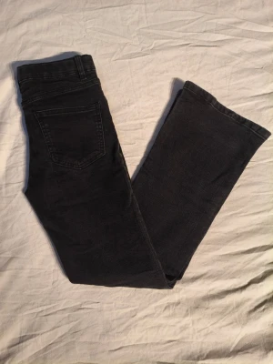 Svarta bootcut jeans - Säljer ett par svarta bootcut jeans i storlek 146 med midwaist och resår med små defekter runt en av bakfickorna.