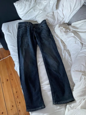 Mörkblå raka jeans från Jack & Jones - Säljer ett par mörkblå jeans från Jack & Jones med raka ben och klassisk femficksmodell. Jeansen har snygga slitningar framtill och en brun läderpatch med logga bak i midjan. Tillverkade i slitstark denim som passar perfekt till en avslappnad stil. Använts 1 gång