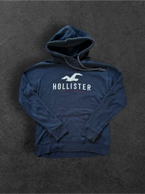 Hollister Hoodie - Mörkblå hoodie från Hollister med stor broderad logga och texten 'Hollister California' på bröstet. Klassisk känguruficka framtill och justerbar dragsko i huvan. Mjuk bomullsblandning och ribbade muddar vid ärmar och nederkant.