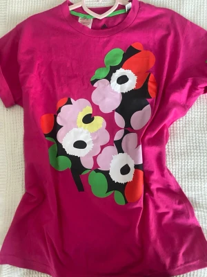 Rosa Adidas x Marimekko t-shirt - Snygg rosa t-shirt från Adidas och Marimekko med ikoniskt blommönster i vitt, svart, grönt, rött och gult på framsidan. T-shirten har rund hals och korta ärmar. Tillverkad i mjuk bomull och har en grön logga på ärmen.