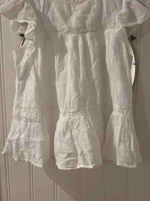 Vit kort volangtshirt i bomull - Söt vit kortklänning med volanger och broderade detaljer. Tillverkad i luftig bomull, perfekt för varma dagar. Klänningen har en avslappnad passform och romantiska detaljer längs ärmar och kjol.