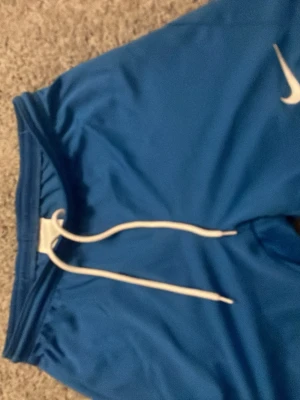 Blå Nike shorts med snörning - Snygga blå shorts från Nike med vit snörning i midjan och elastisk linning. Perfekta för sport eller chill, tillverkade i ett lätt och bekvämt material. Klassisk Nike-logga på benet för en sportig vibe.