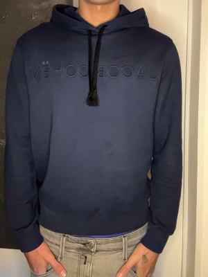 Mörkblå hoodie från Jacob Cohën - Mörkblå hoodie från Jacob Cohën med broderad logga på bröstet och svarta dragsnören i huvan. Klassisk passform med ribbade muddar vid ärmslut och nederkant. Tillverkad i mjukt material som känns skönt mot huden.