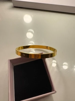 Guldfärgat armband med stenar - Snyggt guldfärgat armband i metall med infällda klara stenar och ingraverade detaljer runt om. Armbandet har en stilren och modern design som passar perfekt till din accessoarsamling.