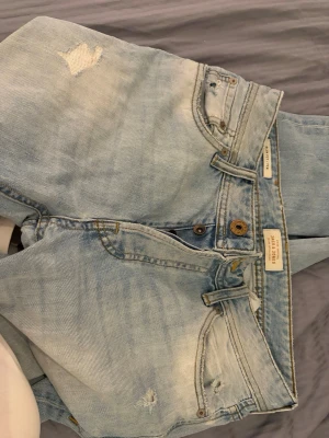 Slim jeans med slitningar från Jack&Jones - Säljer ett par ljusblå jeans från Jack & Jones med slitningar och distressed detaljer. Jeansen har klassisk femficksmodell, knappgylf och slim fit passform. Perfekta för en avslappnad och trendig look.