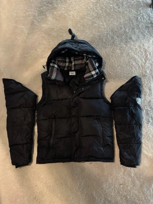 Burberry jacka/väst  - Snygg svart dunväst från Burberry med avtagbara ärmar och huva. Fodrad med klassiskt rutigt mönster i grått, vitt och rött. Två dragkedjefickor framtill och tryckknappar. Perfekt för lager på lager och streetstyle. Ingen return!