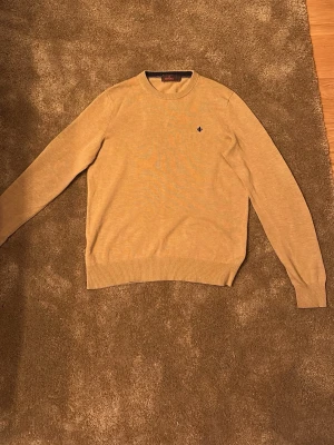 Beige stickad tröja från Morris - Säljer en beige stickad tröja från Morris. med rund halsringning och diskret logga broderad på bröstet. Tröjan har långa ärmar och ribbade muddar vid ärmslut och nederkant. Perfekt för lager på lager och enkel att matcha.