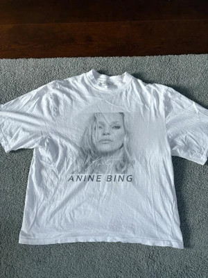 Vit Anine Bing t-shirt med tryck - Säljer en vit t-shirt från Anine Bing med ett stort grafiskt tryck och text på framsidan. T-shirten har rund hals, korta ärmar och en avslappnad passform. Perfekt för dig som gillar stilrena och trendiga plagg.