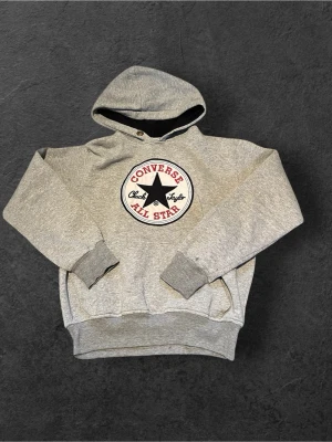 Grå Converse All Star hoodie - Snygg grå hoodie från Converse med det klassiska All Star-märket broderat på bröstet i svart, vitt och rött. Hoodien har huva, ribbade muddar och magficka. Perfekt för dig som gillar streetwear och vill ha en avslappnad stil.