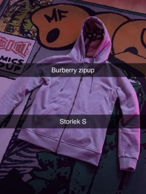 Grå zip hoodie från Burberry - Säljer en grå zip hoodie från Burberry med klassiskt rutigt foder i huvan. Hoodien har dragkedja framtill, två fickor och justerbar huva med snören. Perfekt för dig som gillar stilrena och exklusiva plagg med en twist.