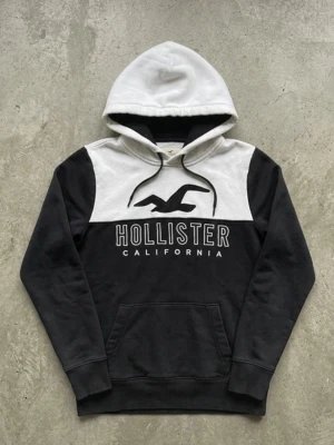 Hollister hoodie - Snygg hoodie från Hollister i svart och vitt med stor logga och texten 'Hollister California' på bröstet. Hoodien har en klassisk huva med dragsko och en stor magficka. Tillverkad i mjukt bomullsmaterial, perfekt för chill dagar.