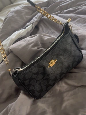 Svart axelremsväska från Coach - Snygg svart handväska från Coach med klassiskt monogrammönster och guldfärgade detaljer. Väskan har en kedjerem i guld, dragkedja upptill och Coach-logga framtill. Tillverkad i läder med en stilren och kompakt form, perfekt för att bära det viktigaste.