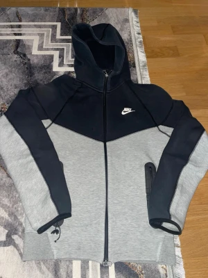 Nike Tech Fleece hoodie svart/grå - Nike Tech Fleece hoodie i svart och grått med dragkedja framtill och vit Nike-logga på bröstet. Hoodien har huva med dragsko, långa ärmar och ficka med dragkedja på sidan. Tillverkad i mjukt och stretchigt material som ger en sportig och clean look.