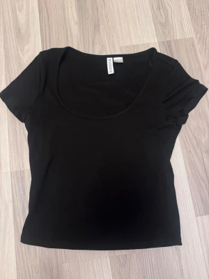 Svart kortärmadd topp från H&M - En enkel och stilren svart topp från H&M med rund halsringning och korta ärmar. Tillverkad i mjuk bomull som känns skön mot huden. Perfekt att matcha med jeans eller kjol för en clean look.